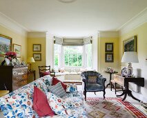 Floral sofa with colour-coordinated scatter cushions, armchair and folding antique table in yellow-painted living room with window bay