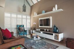 Brown sofa, patterned armchair, curved glass table, sideboard and flatscreen TV on pale brown wall