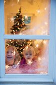 Sisters looking out of window decorated with artificial snow and fairy lights