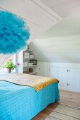 Bett mit blauer Tagesdecke in Dachzimmer mit weisser Holzverkleidung, im Vordergrund blauer Deko Puschel