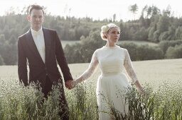 Junges Brautpaar spaziert Händchen haltend durch Haferfeld