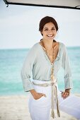 A brunette woman on a beach wearing white trousers and a light tunic with lace detailing