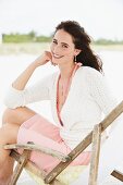 A brunette woman sitting in a deck chair wearing a summer dress and a cardigan