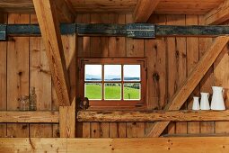 Kleines Sprossenfenster zwischen der Tragkonstruktion einer rustikalen Holzwand, Sammlung weisser Kannen auf einem Balken
