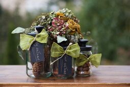 Nuts and pine cones in three storage jars with black lids and labels in front of posy of dried flowers