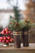 Two small metal buckets decorated with vintage ribbons, spruce twigs and pine cones in front of red apples on cake stand