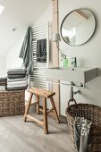 Modern sink below round mirror on wall with rustic wooden stool and wicker basket below