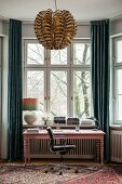 Pink-painted desk in window bay between floor-length curtains and pendant lamp with metallic lampshade in foreground