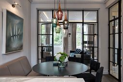 Upholstered bench, black chairs and round table below pendant lamps with glass lampshades of various colours in front of glass partition wall