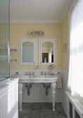Traditional bathroom with vintage-style pedestal sinks against pastel yellow wall and integrated cabinet with mirrored door