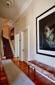 Upholstered bench below portrait of woman on wall and staircase in background in traditional elegant hallway
