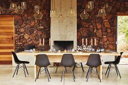Black designer chairs around long solid wooden table in front of rustic stone wall in safari lodge