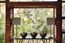 Metal bowls and table lamps arranged on narrow table in front of panoramic window with view of wild landscape