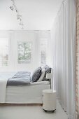 Bett mit grauer Bettwäsche und rundem Rollcontainer als Nachtkästchen vor bodenlangem Vorhang in weißem Schlafzimmer