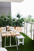 Set table and white bench on balcony with artificial grass floor and small conifers planted in wooden trough