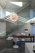 Classic pendant lamp above dining area with white shell chairs and rustic wooden table in front of concrete staircase with glass balustrade