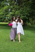 Two barefoot young women walking arm-in-arm across summer meadow, one holding bouquet of roses