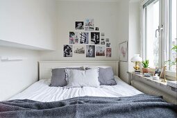 Double bed with grey scatter cushions against white-painted headboard below decorative arrangement of feminine fashion photos on white wall next to window