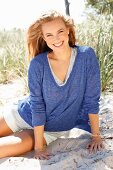 A young woman wearing shorts and a blue mottled shirt sitting on a beach