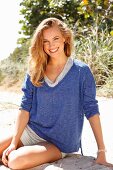 A young woman wearing shorts and a blue mottled shirt sitting on a beach