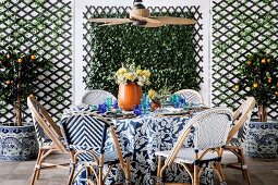 Wicker chairs around a laid table with a floral, blue and white tablecloth under a ceiling fan