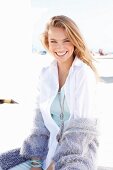 A blonde woman on a beach wearing a white blouse and a purple cardigan