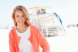 A blonde woman on a beach wearing a white sweater and a salmon coloured jacket