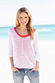 A young woman on a beach wearing a red top, a white open-work jumper and jeans