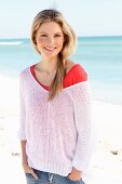 Junge Frau in rotem Shirt, weißem Lochstrickpulli und Jeans am Strand