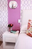 Modern, cubic bedside table below pink glass panel on patterned wallpaper