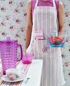 Frau mit gestreifter Küchenschürze vor romantischer Blumentapete hält Glasschale mit Erdbeeren