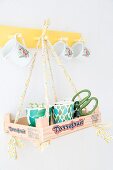 Seedlings in paper cups in recycled wooden crate hung from yellow coat rack by ribbons