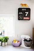 Herbs, fruit basket and mixer on kitchen counter below calendar clock on white-tiled wall