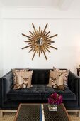 Scatter cushions with stag motifs on black velvet sofa below sunburst mirror on white wall