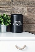 Schwarze Metalldose und Grünpflanze auf Küchenunterschrank vor rustikaler Holzverkleidung
