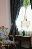 Baroque armchair next to window with heavy blue curtains and printed voile