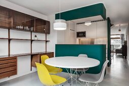 Classic yellow chairs around white table in front of petrol-blue kitchen counter and custom shelving made from exotic wood