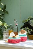 Crocheted watermelon baskets of biscuits next to glasses and swing-top bottles on garden table