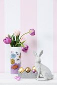 Concrete hare, Easter ornaments and vase of tulips against striped wallpaper