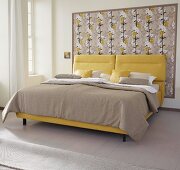 A yellow boxspring bed with a sand-coloured cover against framed Japanese-style wallpaper