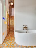 Free-standing modern bathtub against white wall in front of shower area with ornate floor and wall tiles