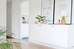 Houseplant, pineapple table lamp and pictures leaning against wall on top of white sideboard in hallway