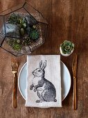 Linen napkin with rabbit motif on place setting with miniature terrarium and succulent in egg cup on wooden table