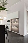 A made-to-measure kitchen cupboard partially built in to a wall with illuminated shelves on a grey wall in an open-plan kitchen