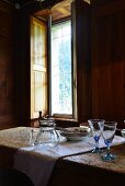 Glasses, carafe and plates on rustic dining table next to window in wood-clad dining room