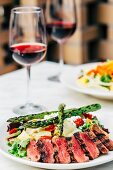 Gegrilltes Rindersteak mit Spargelsalat und Rotweingläsern auf Restauranttisch