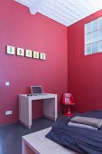 Laptop desk against red wall in minimalist workspace in corner of bedroom with industrial windows