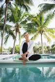 An older woman sitting by a swimming pool under palm trees