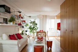 Fitted kitchen, orange stools, white loose-covered sofa and houseplants in one-room apartment