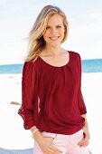 A young blonde woman on a beach wearing a bordeaux red top and light trousers
